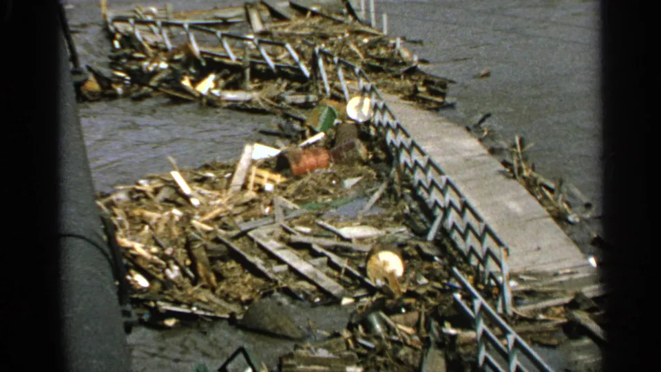 1965 Minnesota home-movie frame showing wreckage, a concrete walkway, and dark river water after flooding.