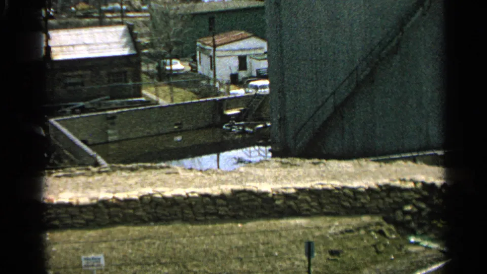 1965 Minnesota 8mm view of a small white riverside building, stone wall, grass, and floodwater nearby.
