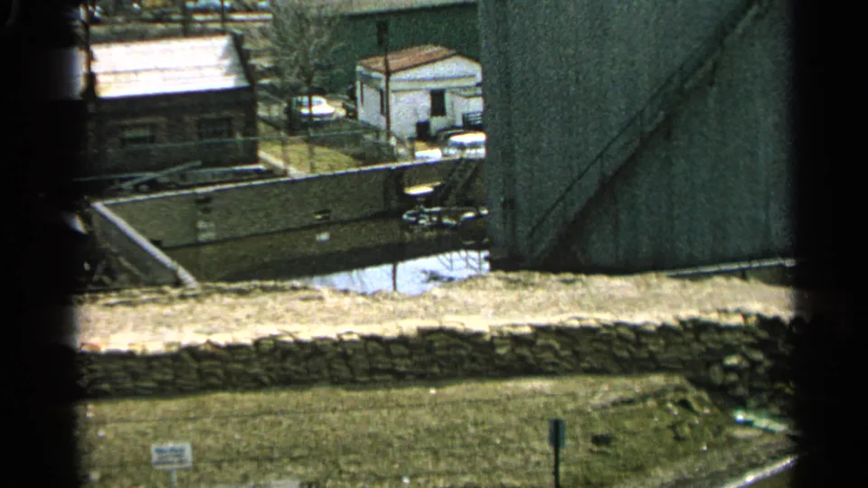 1965 Minnesota frame with a small white building beside water and a stone wall in the foreground.