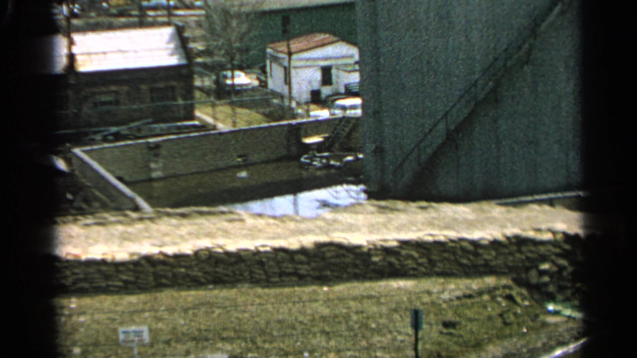 Elevated view of a flooded industrial yard with buildings, retaining walls, and snow patches.