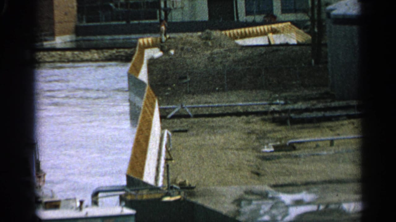 Water beside a muddy yard with pipes, a metal structure, and industrial buildings beyond.