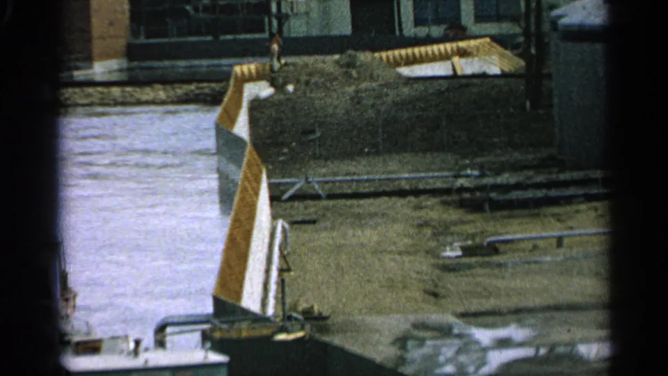 1965 Minnesota 8mm view across water toward a muddy shipping-yard area with pipes, a metal structure, and plain industrial buildings.