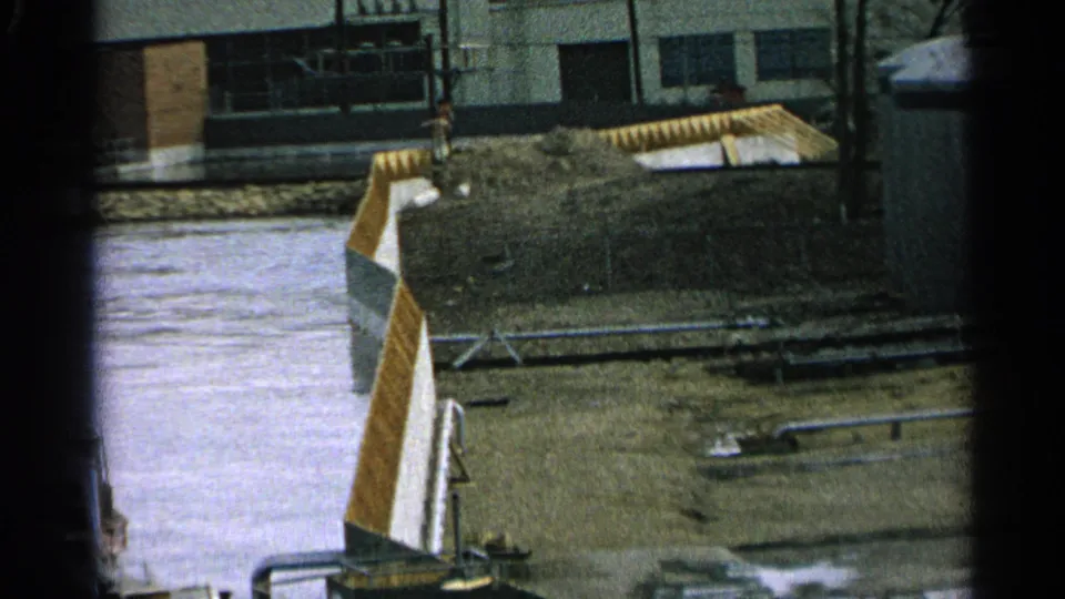 1965 Minnesota home-movie frame with water, muddy ground, pipes, and a yellow-and-white metal structure in a worn industrial yard.