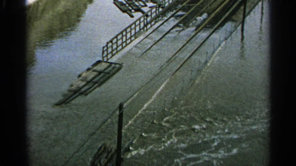 1965 Minnesota 8mm frame showing a flooded industrial surface with metal rails, water movement, and a small distant person near the lower area.