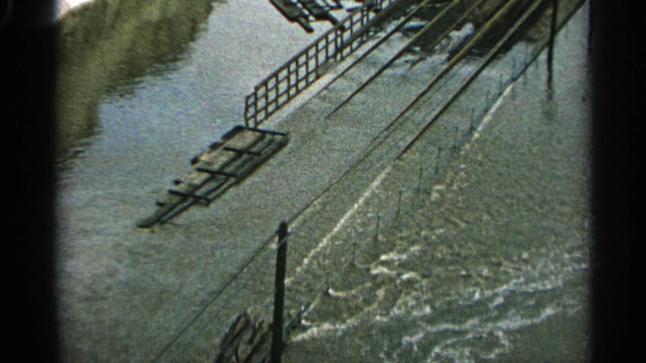 1965 Minnesota 8mm frame showing floodwater moving through an industrial area with railings and submerged track-like elements.