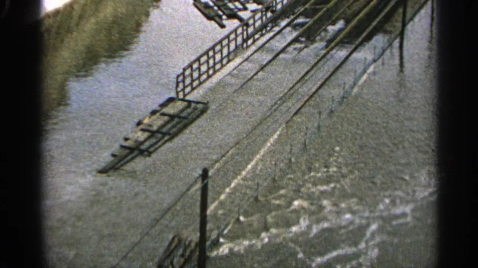 Minnesota 1965 home-movie frame showing floodwater over industrial ground, with railings and track-like lines still visible.