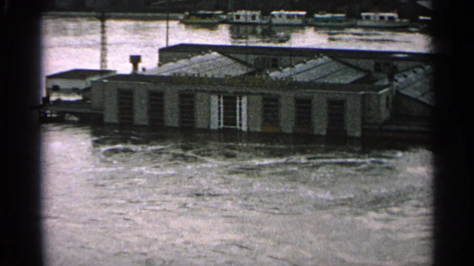 1965 Minnesota 8mm frame showing a low riverside building surrounded by floodwater, with distant boats or structures behind it.
