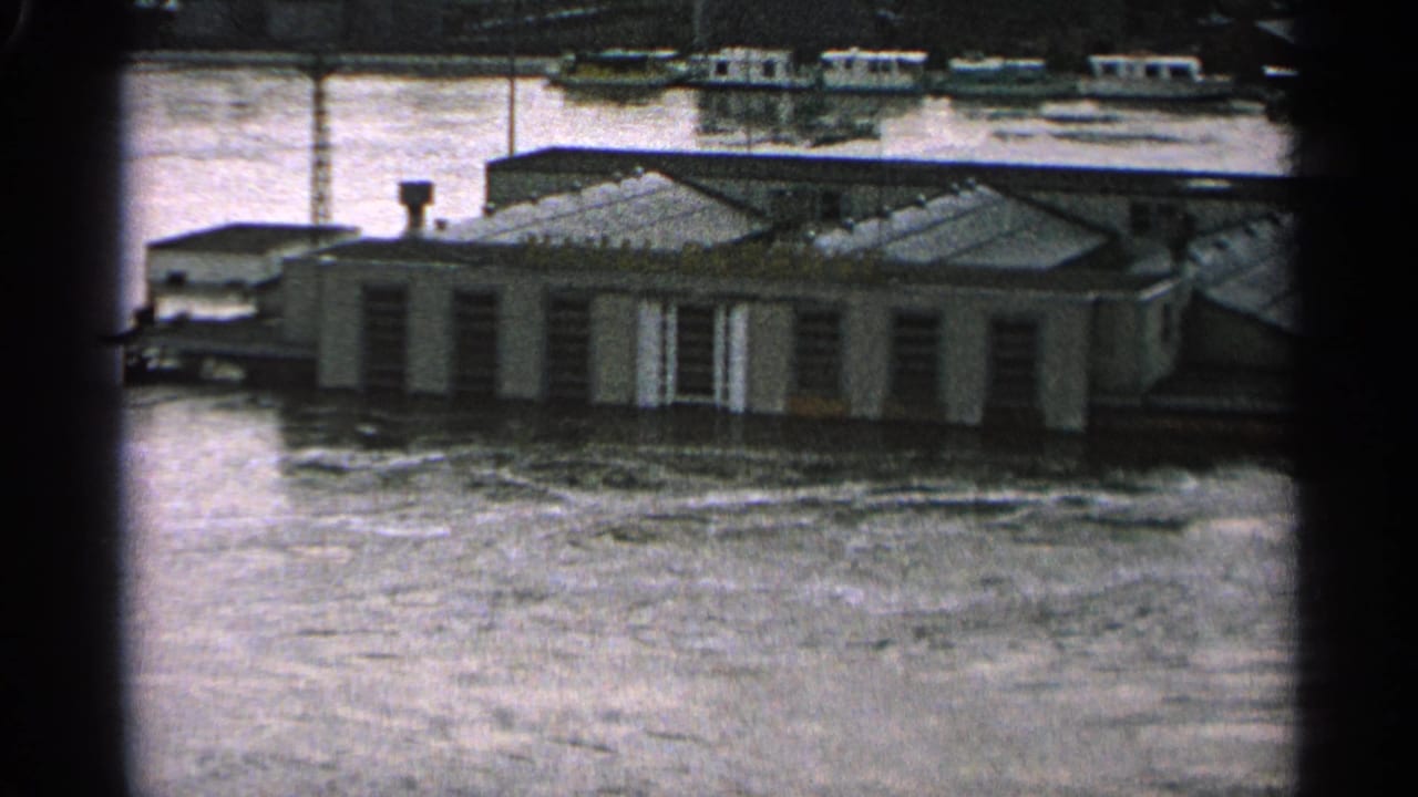 Floodwater surrounds a low building with small boats visible in the background.