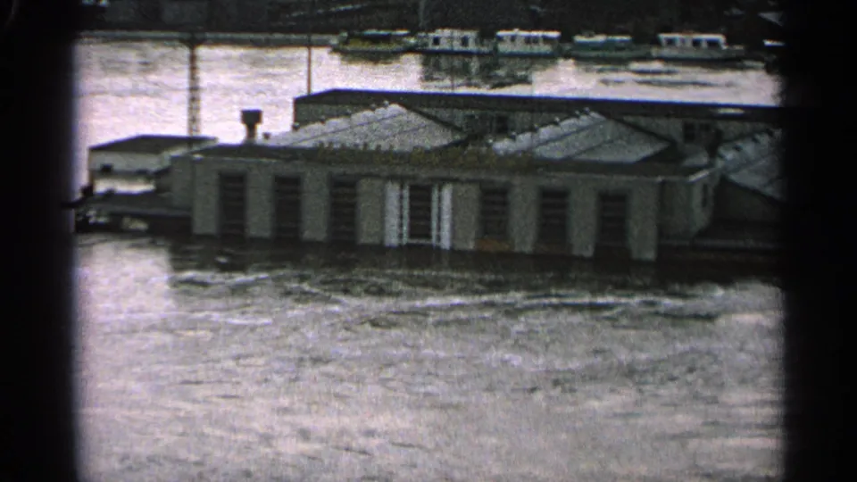 1965 Minnesota 8mm frame showing floodwater around a low riverside building, with small boats beyond the structure.