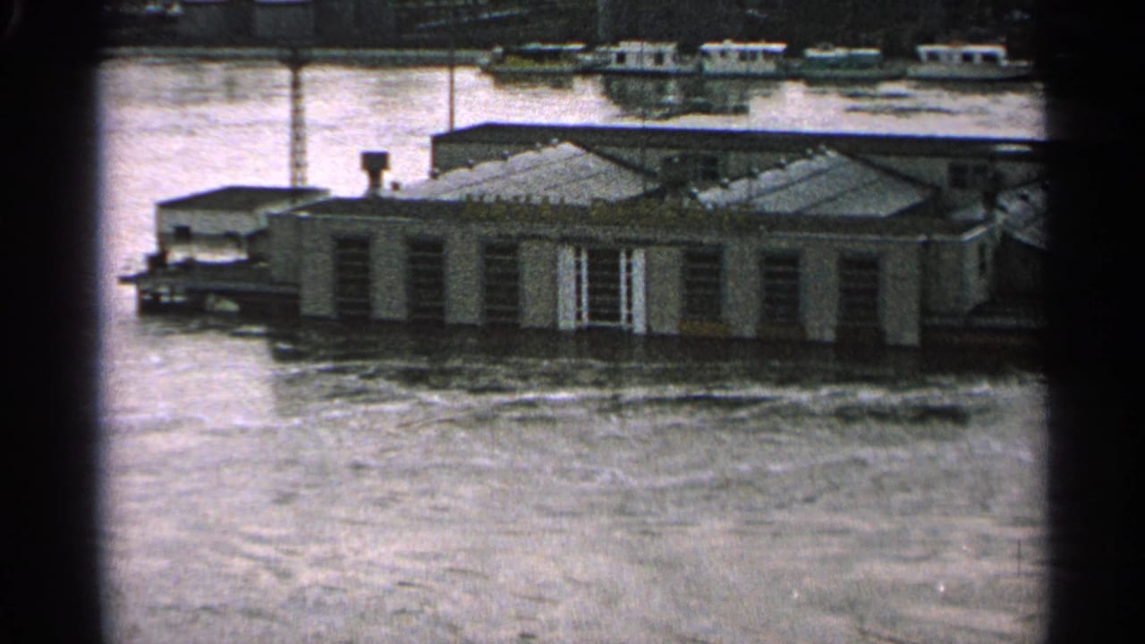1965 Minnesota flood footage shows a partly submerged building and boats beyond, seen through soft, grainy 8mm film.