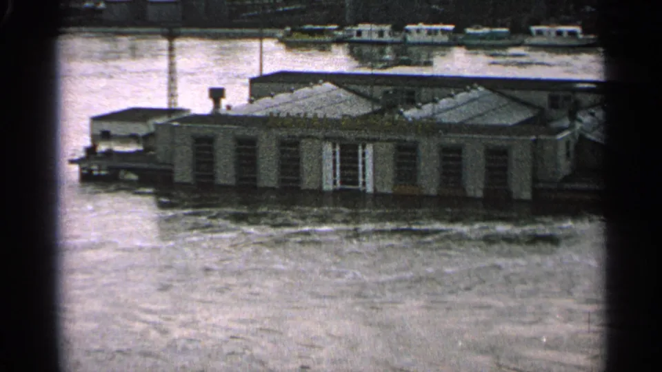 1965 Minnesota flood footage shows a partly submerged building and boats beyond, seen through soft, grainy 8mm film.