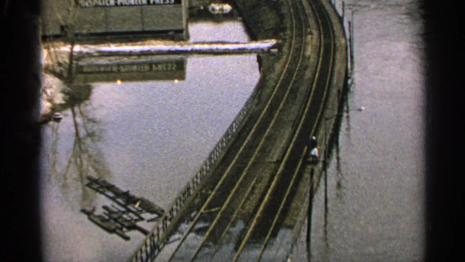 Elevated 1965 Minnesota view of floodwater beside rail tracks, with a partially submerged Dispatch-Pioneer Press sign and scattered debris.
