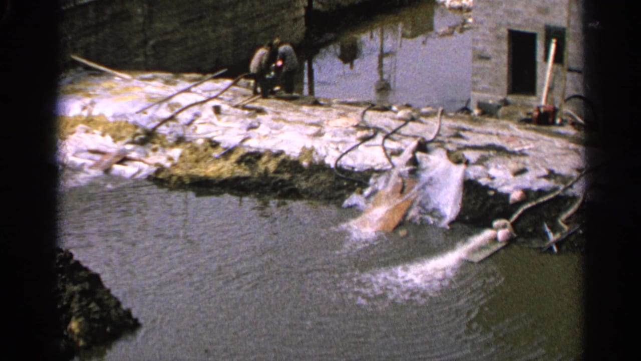 Drainage pipe sprays water into a pond beside a muddy worksite and masonry building.