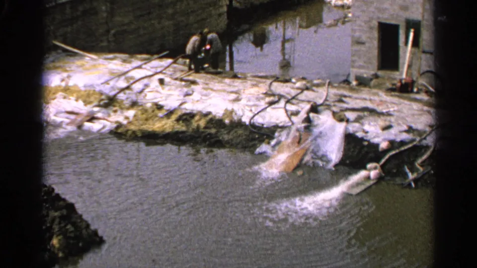 1965 Minnesota 8mm frame showing water discharging from a pipe into a pond while two distant people stand near a masonry wall.
