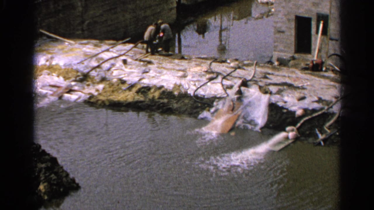 Water continues pouring into the pond near a shoreline worksite with two people in the background.