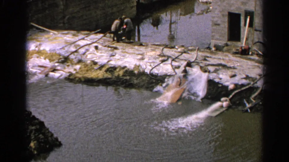 1965 Minnesota home-movie frame of a muddy pond edge, pipe discharge, masonry wall, and two distant observers at a worksite.