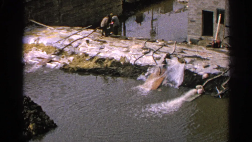 1965 Minnesota 8mm frame showing the drainage outlet, pond ripples, scattered hoses or rails, and two distant people near a building.
