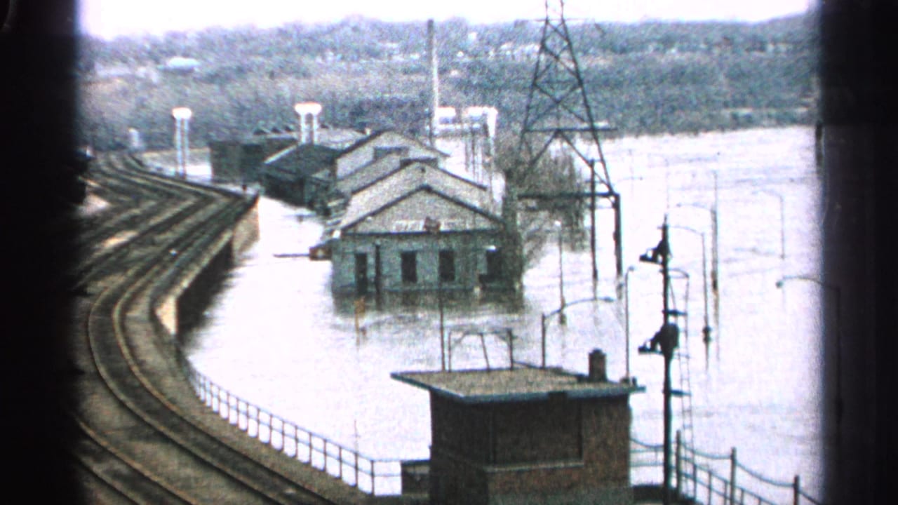 Floodwater surrounds buildings beside curved train tracks and utility structures.