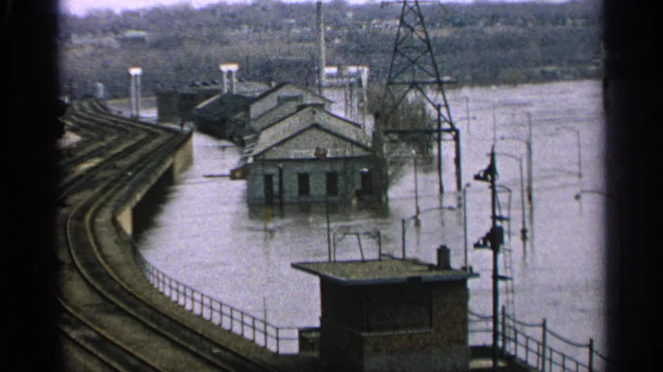 1965 Minnesota flood footage with buildings partly underwater beside curved railroad tracks and visible utility infrastructure.