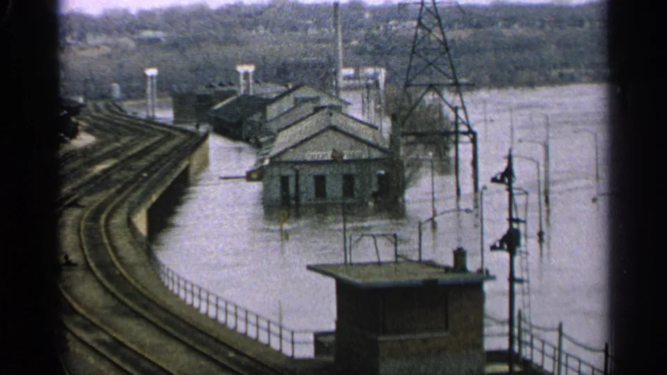 1965 Minnesota rail-side flood scene with murky water around buildings, track curves, poles, and a small shelter in frame.
