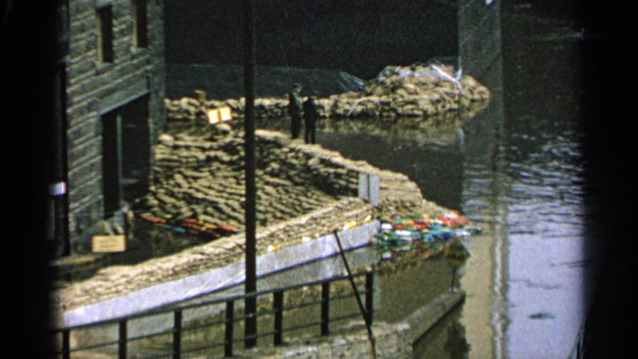 1965 Minnesota flood scene with stacked sandbags protecting a waterfront building as water rises nearby.