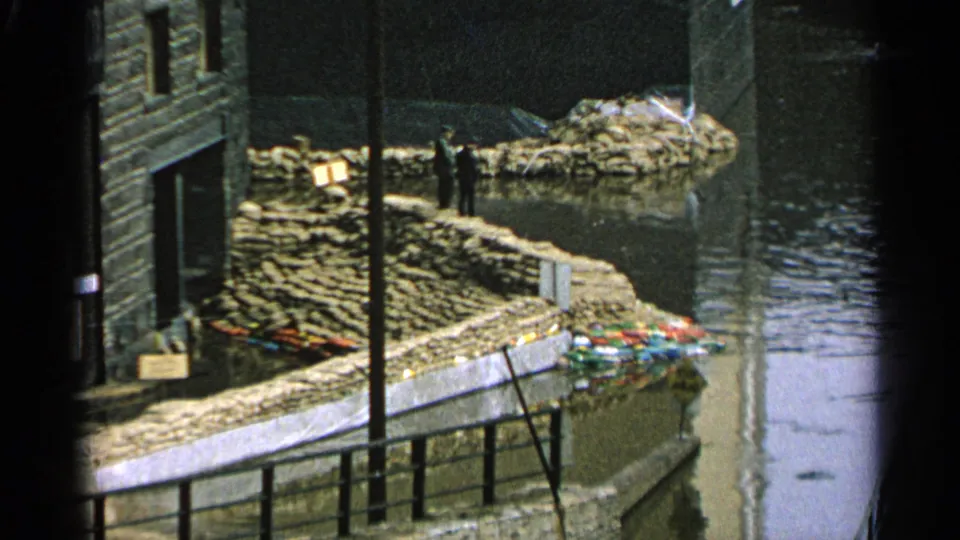 1965 Minnesota flood scene with stacked sandbags protecting a waterfront building as water rises nearby.