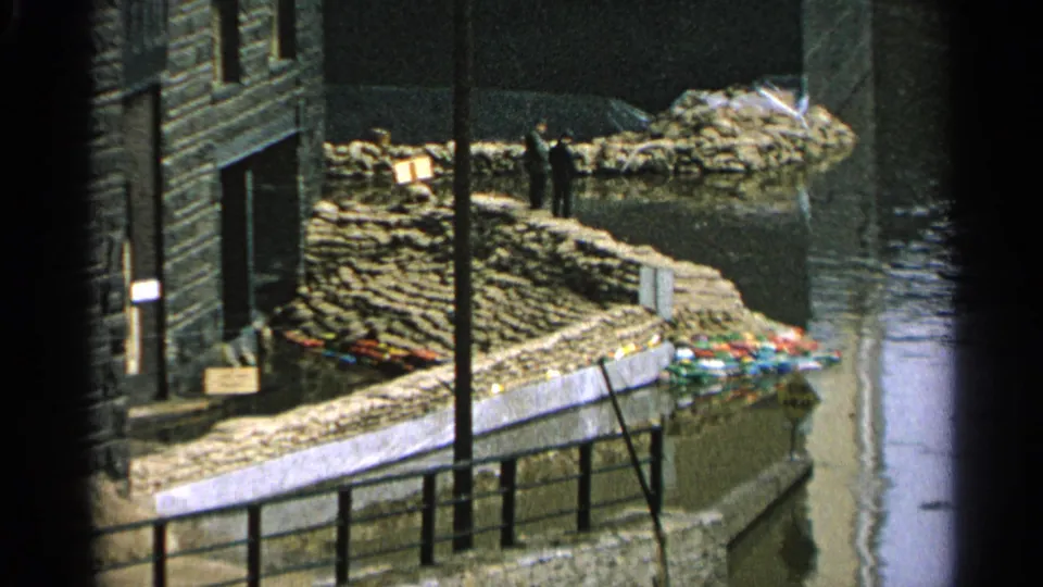 1965 Minnesota home-movie view of a sandbagged waterfront building with a person standing near the flood barrier.