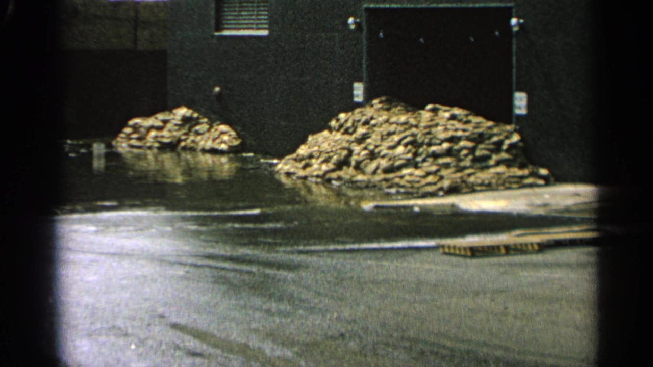 Flooded industrial yard with pooled water, rubble piles, doors, and dark building wall.