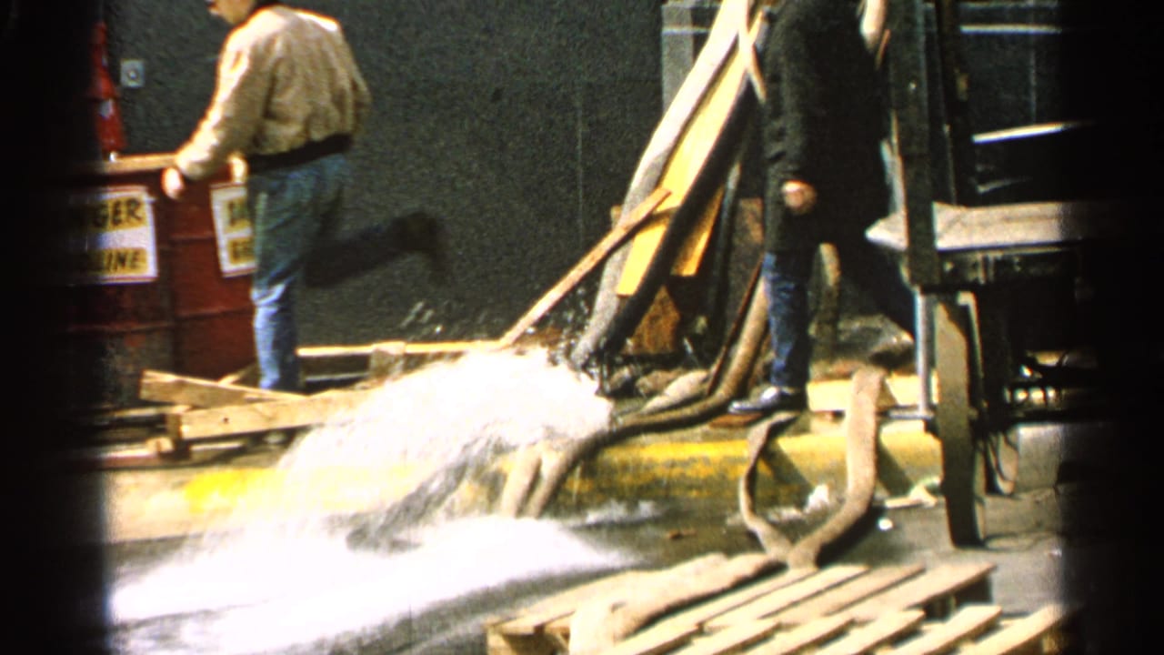 Two adult men near red barrels as water gushes from a hose onto wet pavement.
