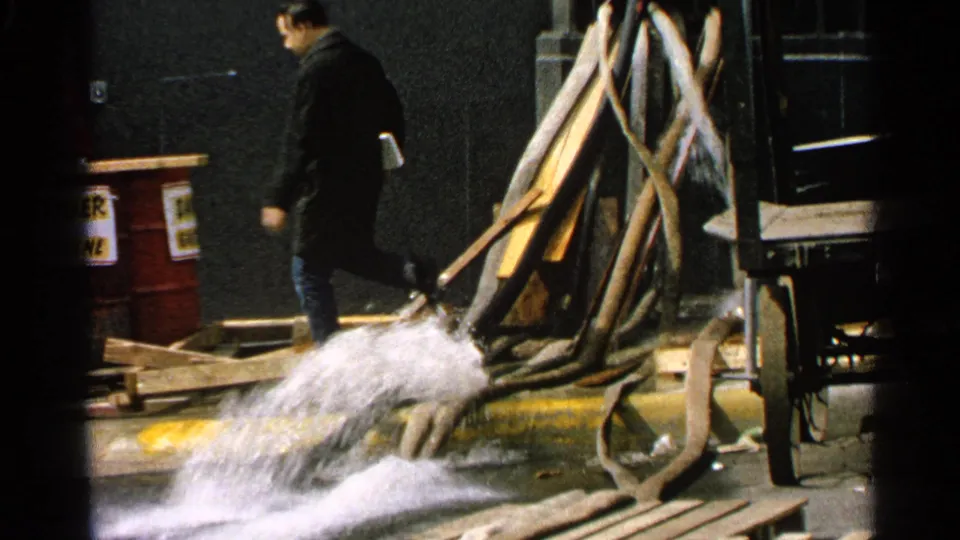 1965 Minnesota 8mm frame of an adult man walking near a forceful hose spray in an industrial yard.