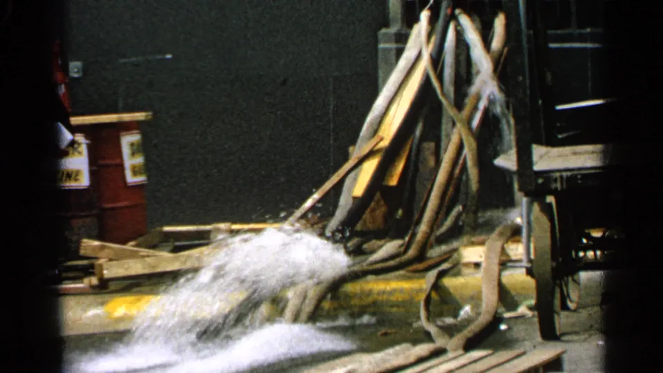1965 Minnesota 8mm frame showing water pouring from a hose near red barrels, lumber, and an industrial vehicle edge.