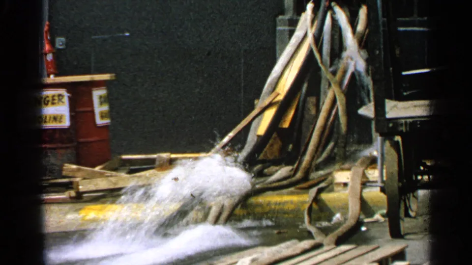 1965 Minnesota 8mm frame of a forceful hose stream crossing a dock area with barrels, hoses, boards, and wet pavement.