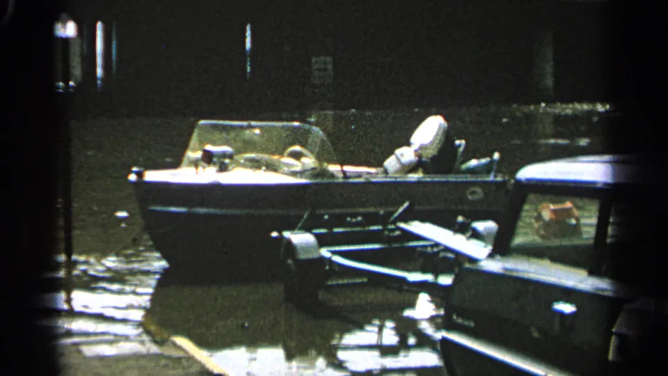 The 1965 Minnesota dock scene shows a small boat, trailer frame, and vehicle beside standing water and dark waterfront structures.