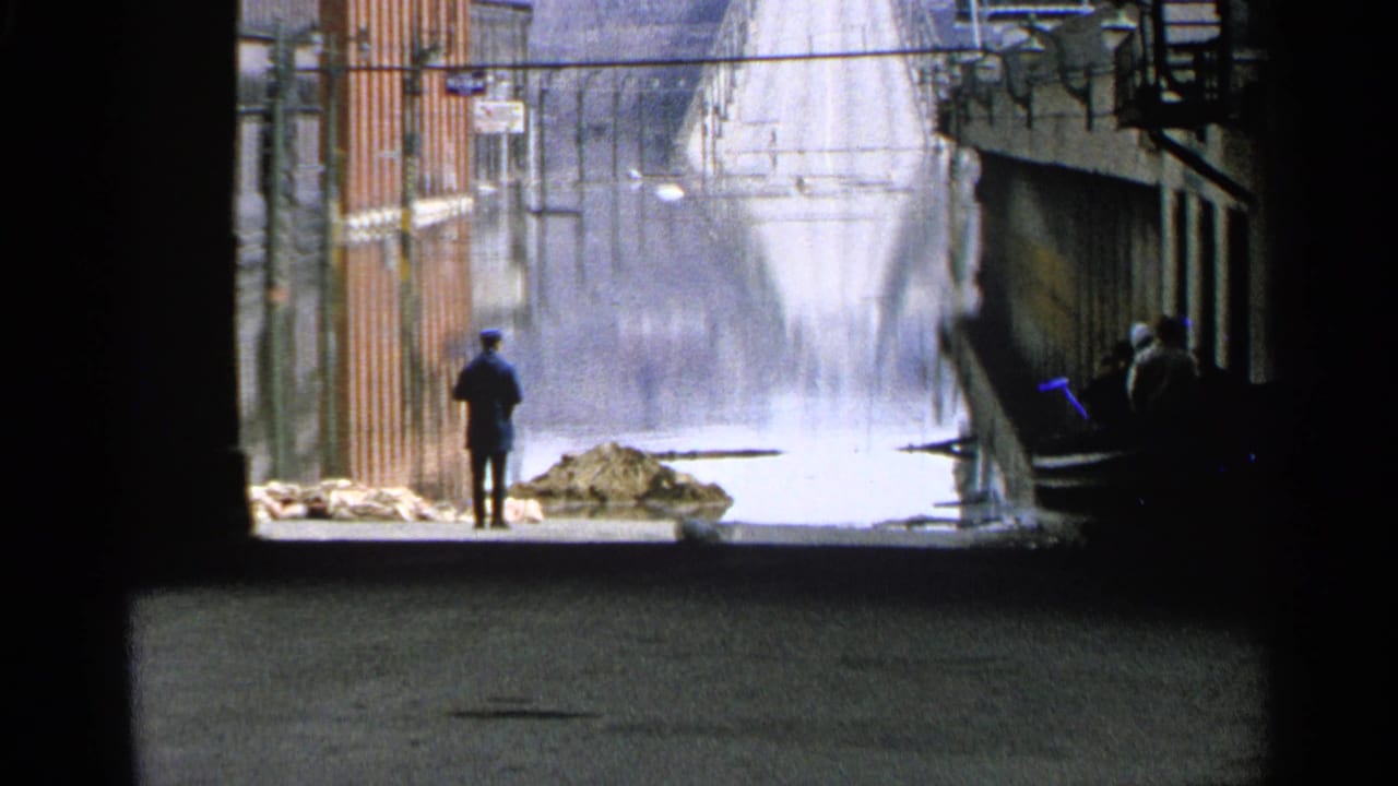 A distant man in dark clothing observes a flooded Minnesota street with sandbags and storefronts in this 1965 8mm frame.