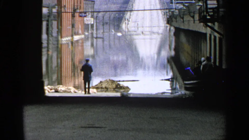 A distant man in dark clothing observes a flooded Minnesota street with sandbags and storefronts in this 1965 8mm frame.