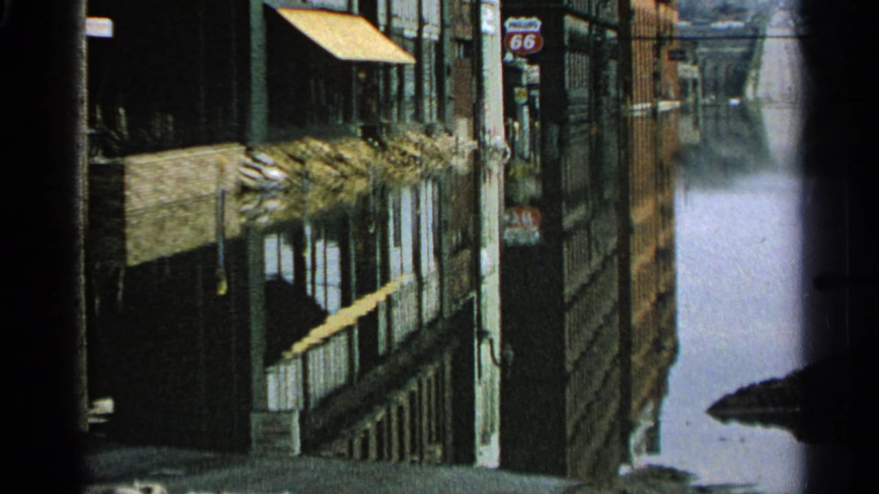 Floodwater reflects buildings, awnings, utility poles, and a visible 66 road sign along a Minnesota commercial street in 1965 footage.