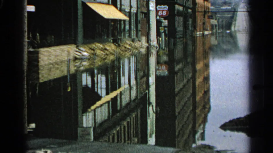 Floodwater reflects buildings, awnings, utility poles, and a visible 66 road sign along a Minnesota commercial street in 1965 footage.