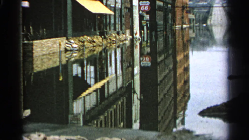 The 1965 home-movie frame looks down a flooded Minnesota street with awnings, utility poles, building fronts, and reflective standing water.