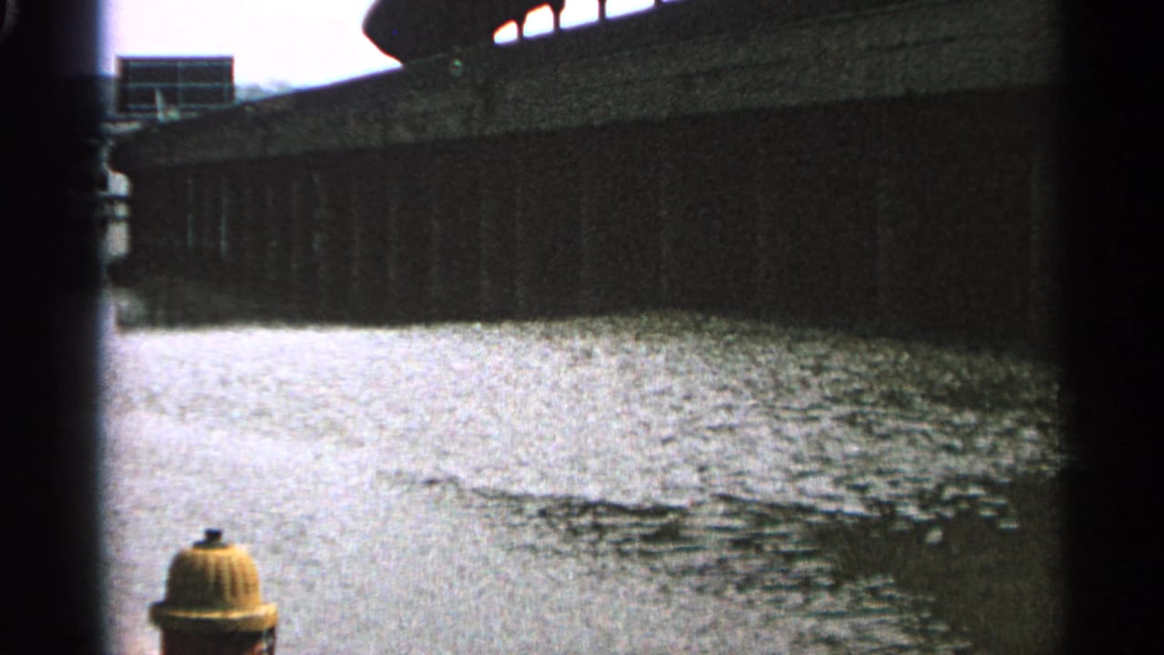 1965 Minnesota 8mm frame showing rippling water beneath a concrete bridge or wall, with a yellow fire hydrant at the lower edge.
