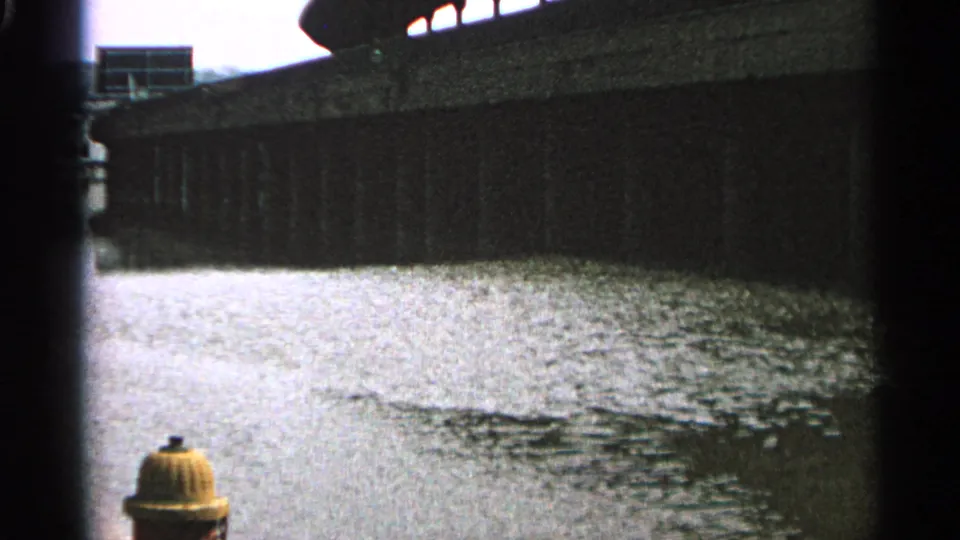 1965 Minnesota 8mm frame showing rippling water beneath a concrete bridge or wall, with a yellow fire hydrant at the lower edge.