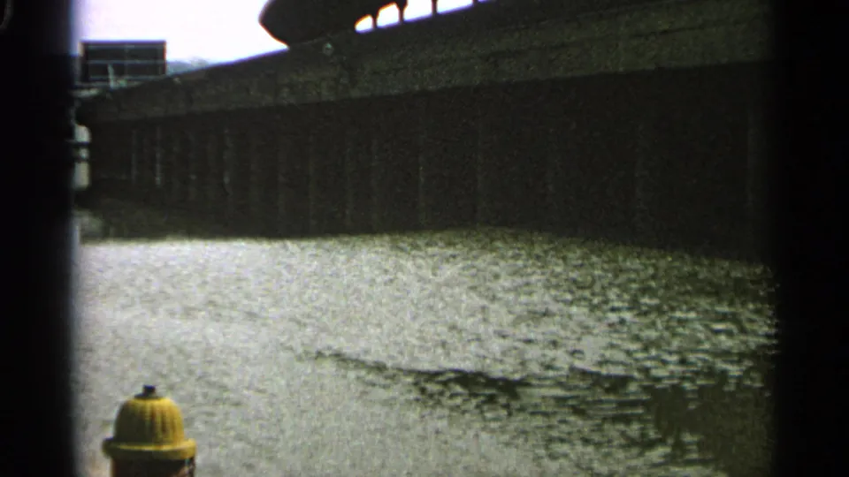 1965 Minnesota 8mm view of water flowing below a concrete bridge or retaining wall, framed by dark film edges and a hydrant below.