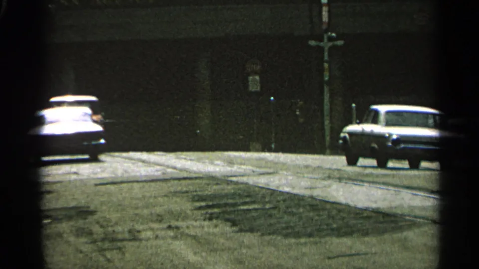 1965 Minnesota street view with mid-century cars moving along a paved road beside a dark wall and utility pole.