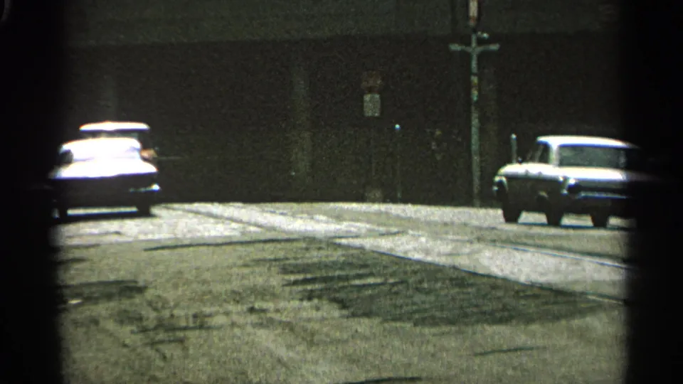 1965 Minnesota 8mm street frame showing two cars on the roadway, a dark wall behind them, and utility poles near the curb.