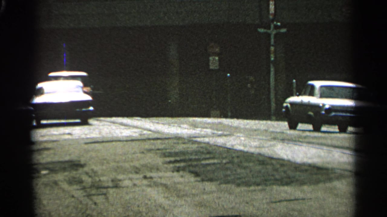 1965 Minnesota 8mm frame showing cars on a quiet street beside a dark building wall, with soft grain and partial framing.