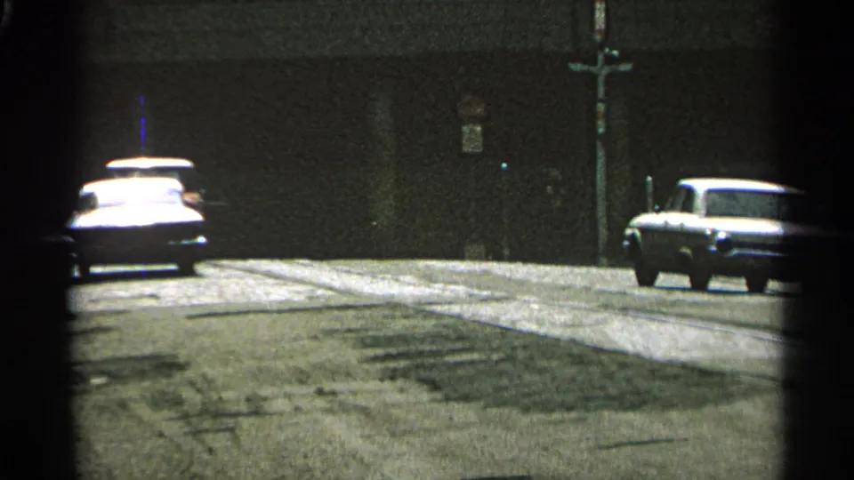 1965 Minnesota 8mm frame showing cars on a quiet street beside a dark building wall, with soft grain and partial framing.