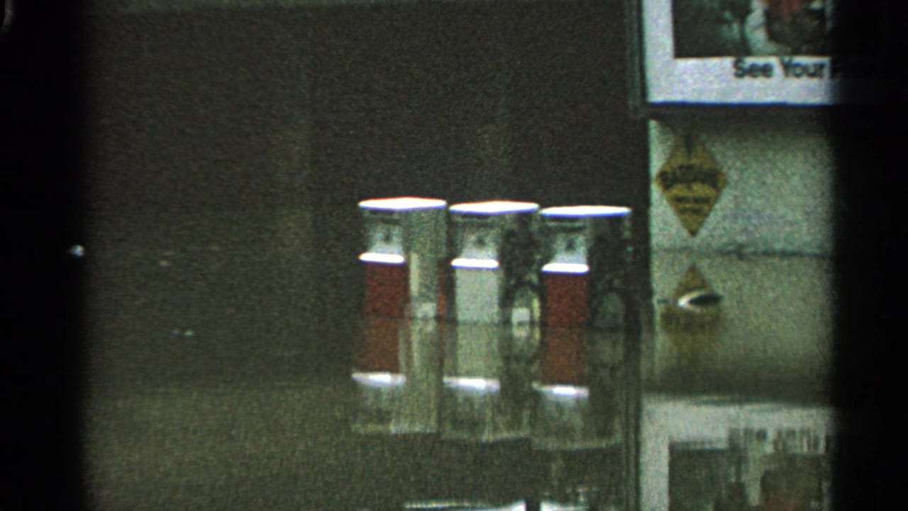 Service-station detail with red-and-white pump canisters, wall signs, and a partially visible poster in 1965 Minnesota footage.