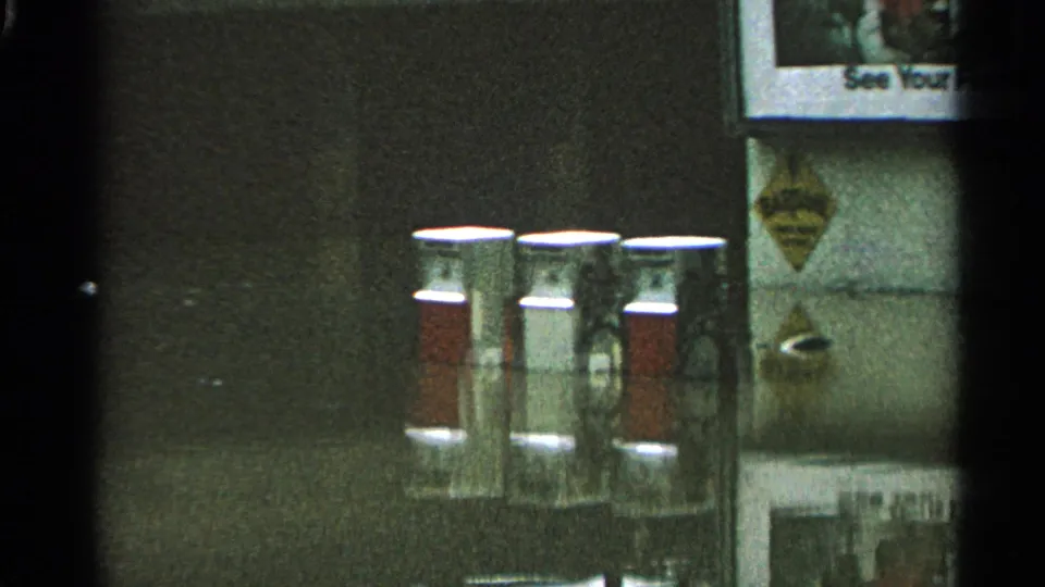 Service-station detail with red-and-white pump canisters, wall signs, and a partially visible poster in 1965 Minnesota footage.