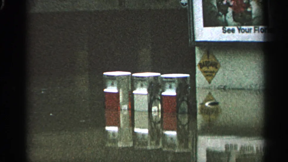 Close 8mm view of red-and-white gas pump canisters and adjacent wall signs, likely from a local service-station setting.