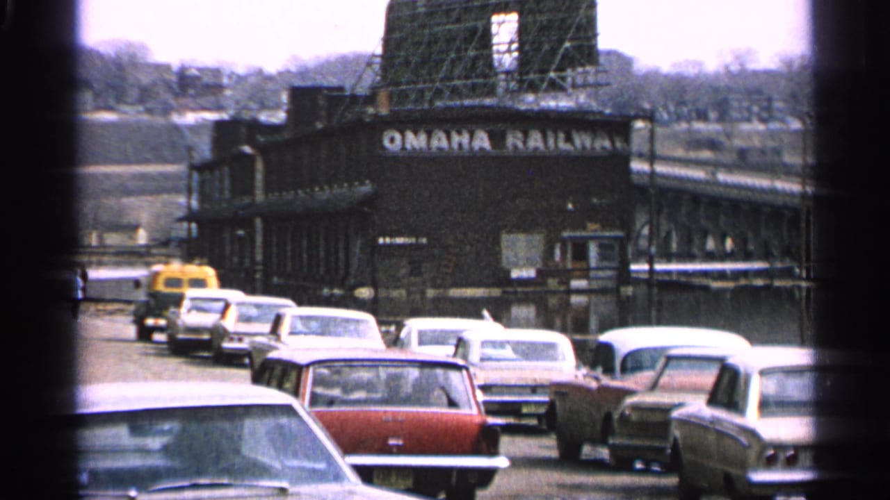 Traffic passes an Omaha Railway building with classic cars and a bridge in the background.