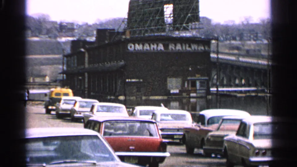 1965 Minnesota home-movie frame showing classic cars on a street beside an Omaha Railway building and bridge structure.